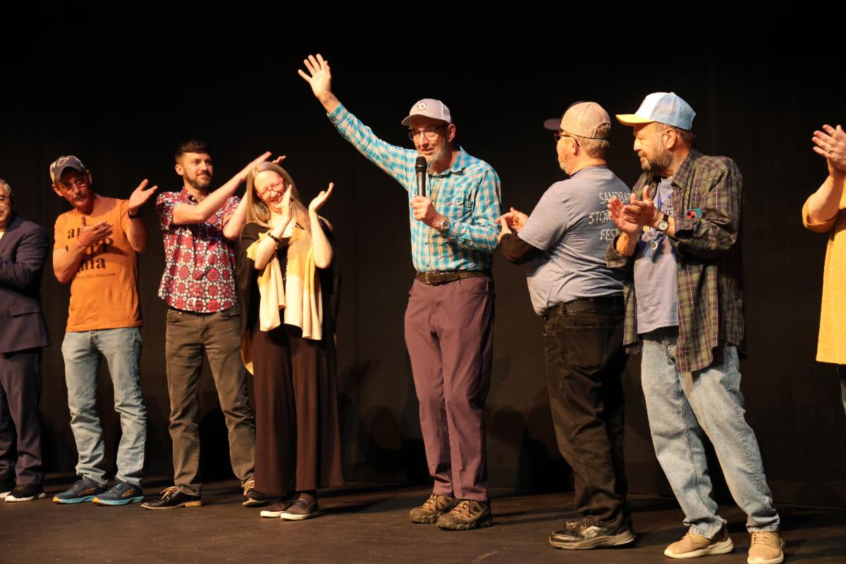 On stage with a black curtain background stands nine people clapping for the presenter in the middle.