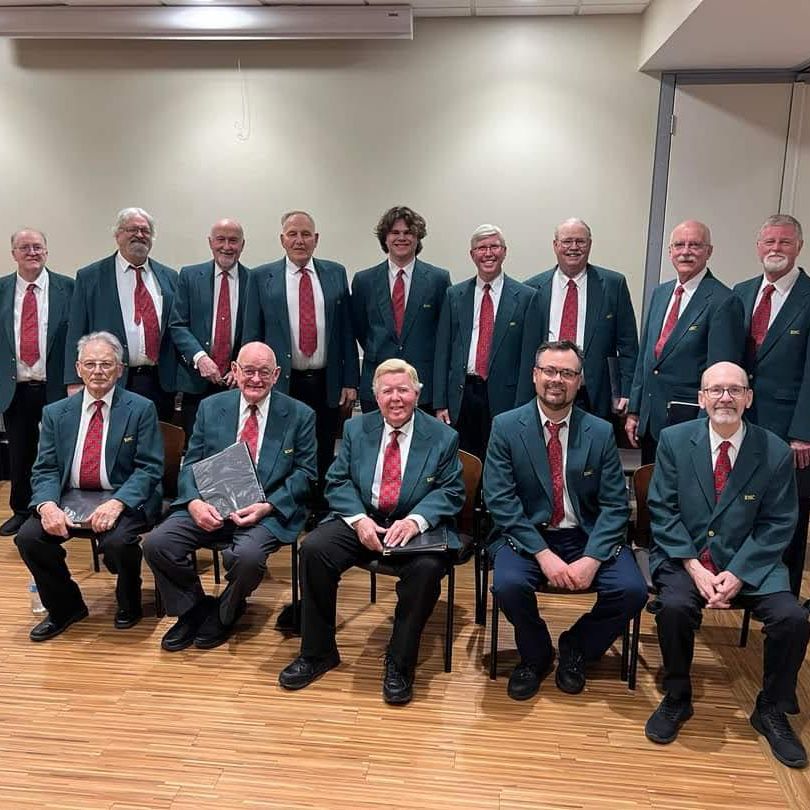 Alt text: Group photo of the Rochester Male Chorus. The ensemble is arranged in two rows - one standing and one seated - in what appears to be a rehearsal or performance space. The members are wearing matching formal attire consisting of dark green blazers, white shirts, and red patterned ties. The group includes approximately 16 men of various ages, with some members holding what look like music folders. They are posed professionally against a neutral wall background with a wooden floor beneath them.