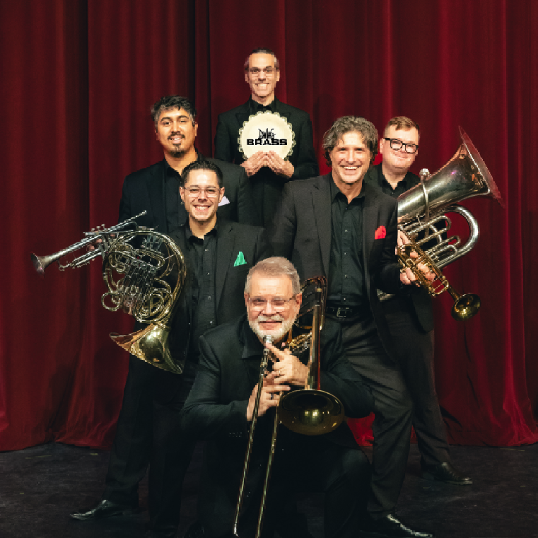 A promotional photo of the Dallas Brass ensemble against a deep red curtain backdrop. The six musicians are dressed in black formal attire with colored pocket squares. They are arranged in a staged pose with their brass instruments including French horn, trombone, and trumpet. One member in the center holds a 'Dallas Brass' sign. The lighting is theatrical and professional.