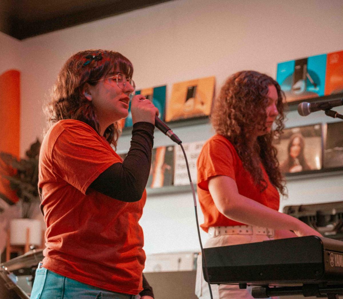 The image shows two young performers in matching bright orange t-shirts on a stage. On the left, a person with glasses and shoulder-length wavy brown hair is speaking or singing into a microphone. They're wearing a long-sleeved black shirt under their orange t-shirt and appear to be fully engaged in their performance. On the right, another performer with long curly hair stands next to an electronic keyboard or synthesizer. The background shows what appears to be an indoor performance space with colorful artwork or panels displayed on the wall behind them.