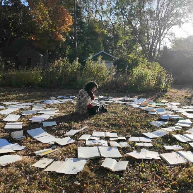 A person sits cross-legged in the center of a large outdoor installation of hundreds of open books scattered across a grassy field. The books are spread in a circular pattern around the seated figure who appears to be reading. The scene is set in autumn with trees displaying fall foliage in the background and bright sunlight creating lens flare effects. A green house is visible among the trees in the distance. The artistic installation creates a surreal library setting in nature.