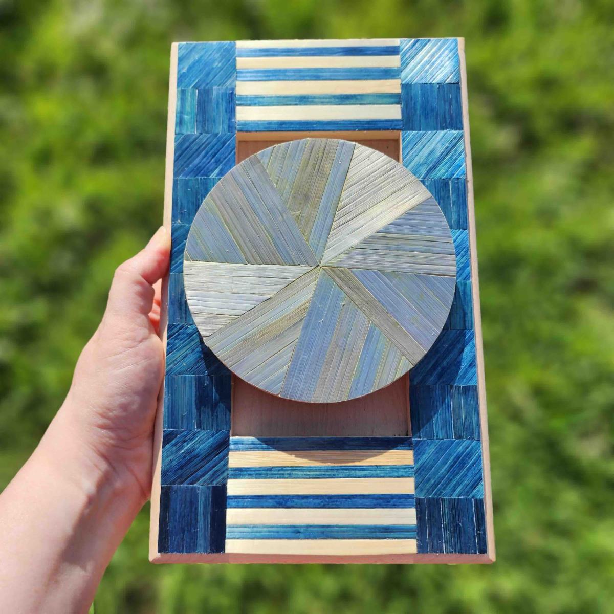 A hand holds a rectangular decorative art piece featuring French straw marquetry techniques. The artwork displays a geometric design with blue and natural colored straw strips arranged in various patterns. The center features a circular medallion with radiating straw pieces creating a sunburst or flower pattern. The surrounding border combines blue straw sections with natural cream colored horizontal stripes and corner elements. The piece demonstrates the intricate craftsmanship of traditional straw marquetry with its precise geometric arrangements and contrasting colors. Green foliage is visible in the blurred background.