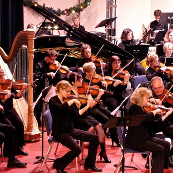 The image shows an orchestra or chamber ensemble performing in what appears to be a concert hall or event venue. In the foreground, there are string musicians (violinists and violists) dressed in formal black attire playing their instruments while seated. A harp is visible on the left side of the image. Behind the string section, there appears to be a grand piano with its lid raised, and other orchestra members including what seem to be wind or brass instrumentalists. The setting has subtle holiday decorations visible, possibly garland, suggesting this might be a seasonal or holiday concert. The lighting gives the scene a warm, atmospheric quality typical of formal classical music performances.