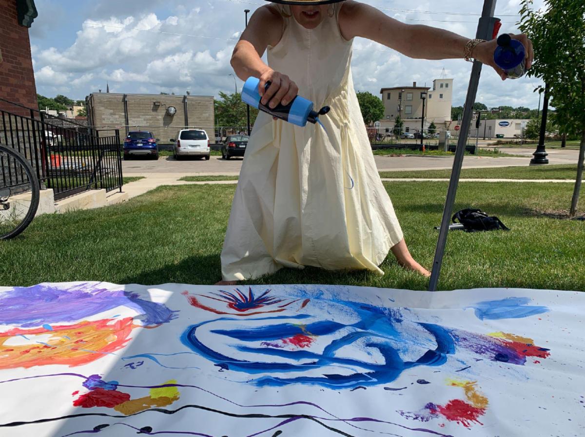A person in a cream-colored sundress and sun hat is creating colorful art outdoors. They're squeezing blue paint from a bottle onto a large white canvas or paper laid on the ground. The canvas already has vibrant red, blue, purple, and yellow paint markings. The scene takes place in a grassy area with buildings and parked cars visible in the background on a sunny day