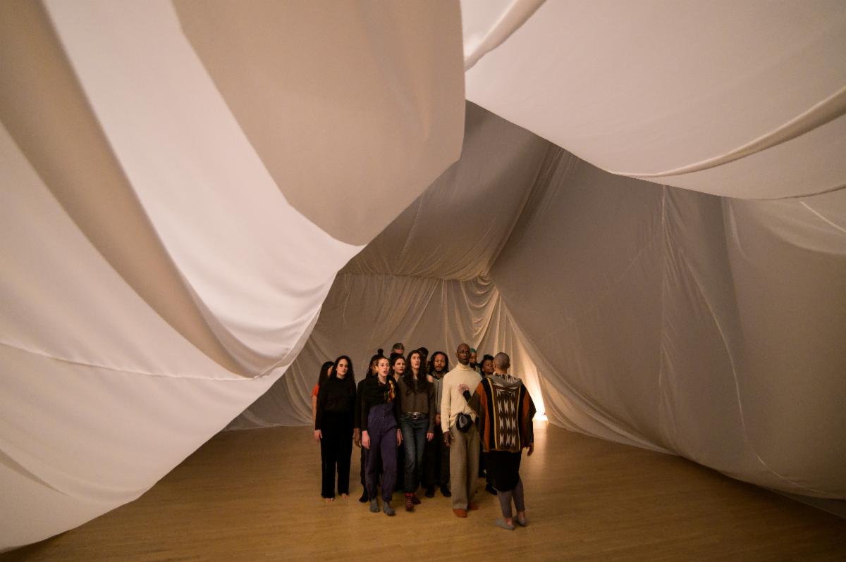 A cluster of people stand inside a huge, lofty white tent. The billowy drapes of the tent fold and crease in many places. A bright light shines through the opening of the tent, illuminating the hardwood floor. The group faces us, except for one person