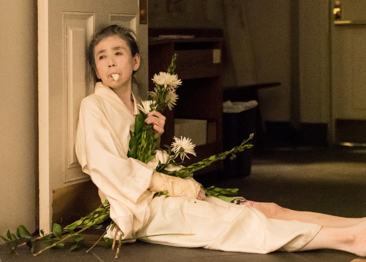 Eiko slumps against an open door. She wears a white kimono and cast on her arm and holds a bouquet of tall white flowers with leafy stalks. Some of the flower’s white petals pour out of her closed mouth. She looks out the open door. 

