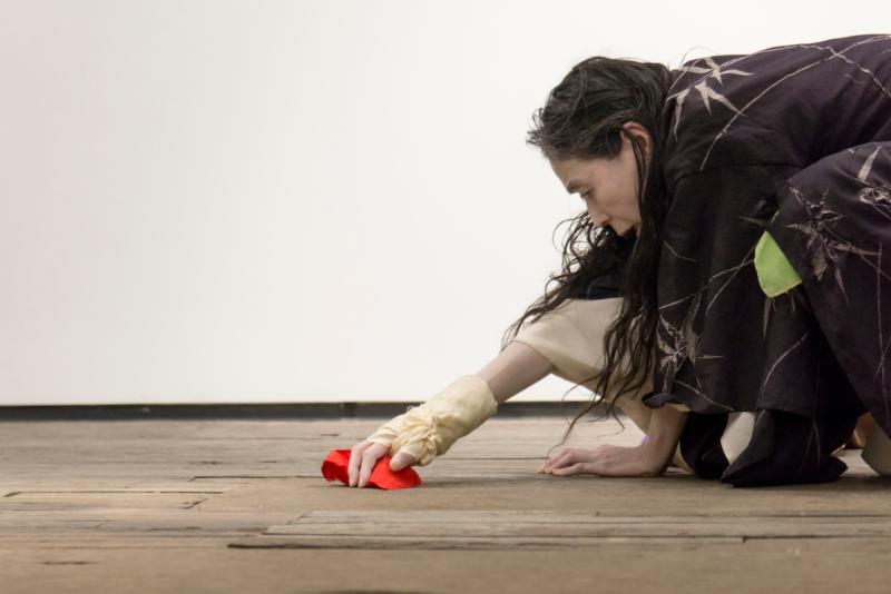 Eiko kneels into a wooden floor. Her arm is stretched out in front of her, covered in a yellow cast. She holds a bright red object. Her other hand grips the floorboards, he knuckles buckling. Her long black hair dangles beneath her. 
