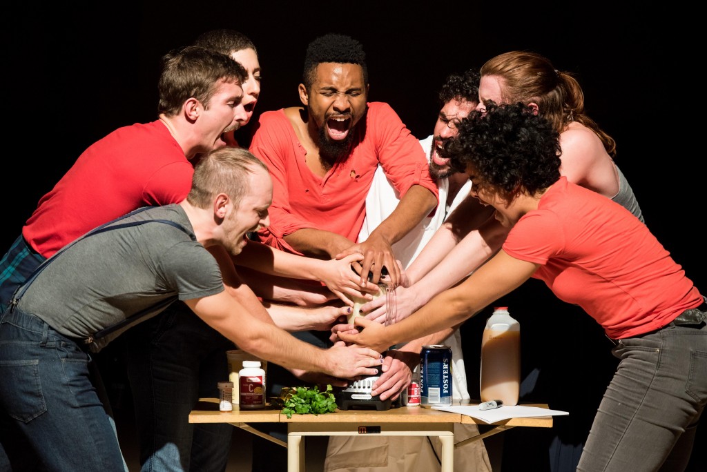 A group of brightly lit people stand huddled together. Their mouths are wide open, perhaps singing or yelling. They grip a blender, filled with beige liquid. 