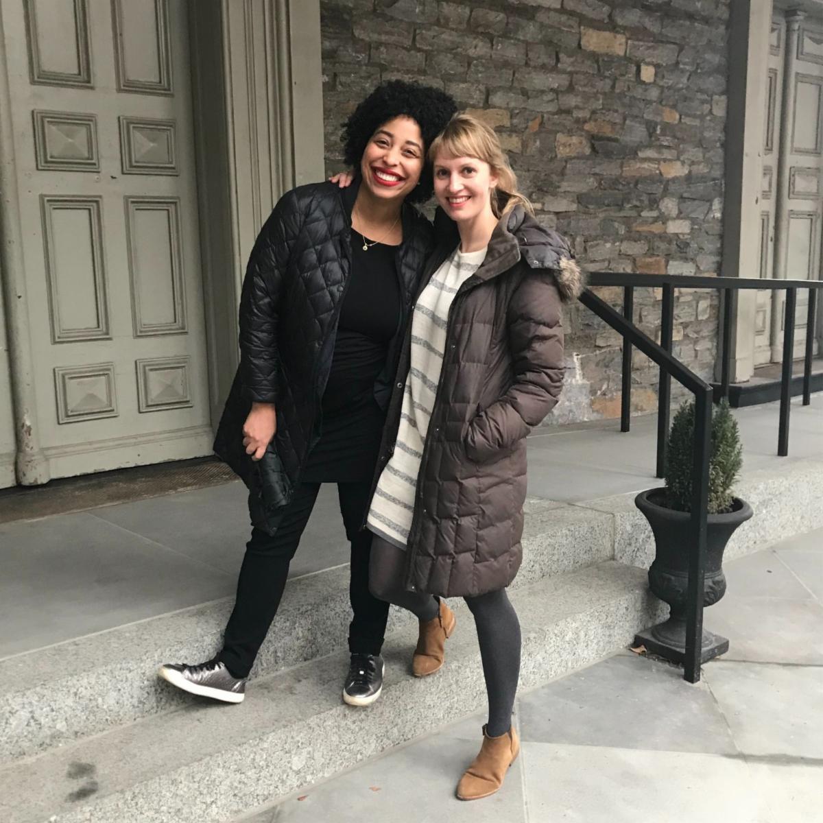 Kristin and Lydia are smiling and sharing a tender side hug. They stand on tall stone steps in front of the entrance to a building. The building’s facade is composed of grey bricks and large grey wooden doors. They both wear dark winter coats. 
