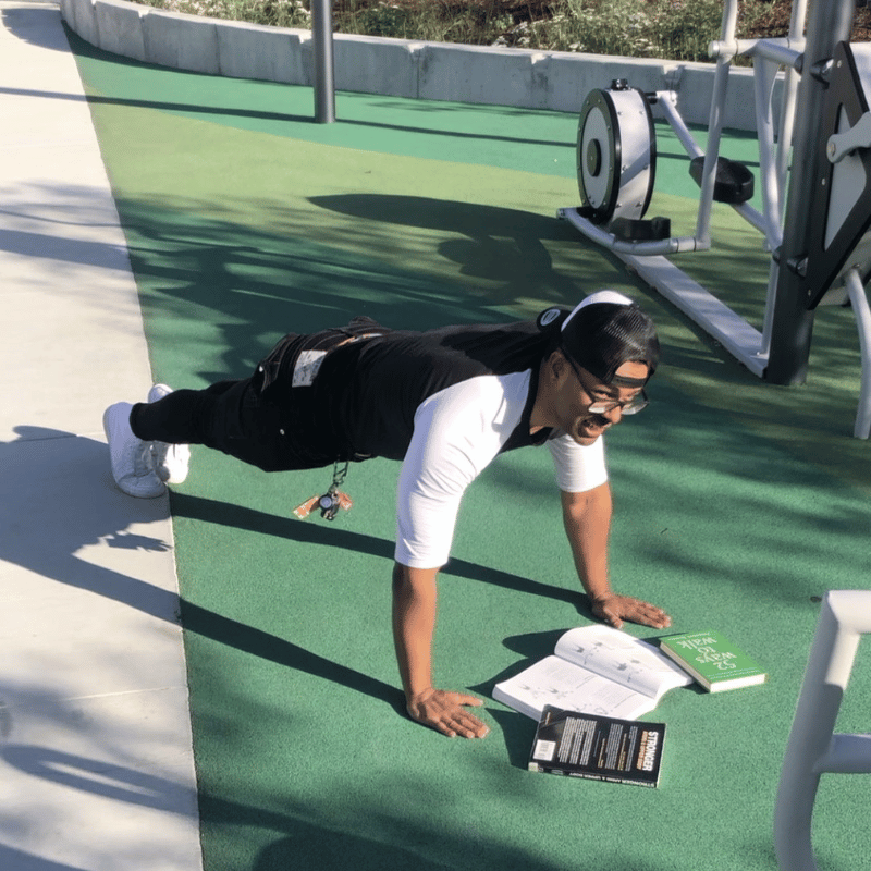 A library worker poised to do pushups outside the library near the exercise equipment, with books laid out underneath him.