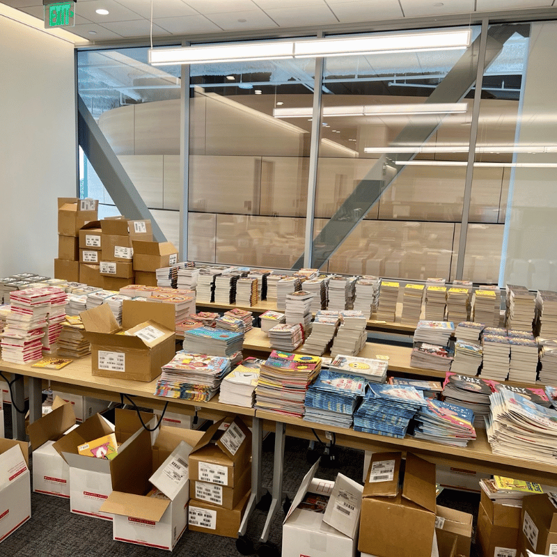 Piles of books, organized by title and age, laid out on tables in the library office in preparation for Summer Learning Challenge.