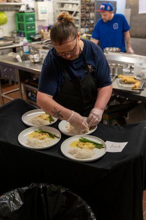 A Talonâs Table chef's stands at a well-lit counter, carefully plating a meal with focused attention to detail, arranging food neatly on a clean white plate for presentation.