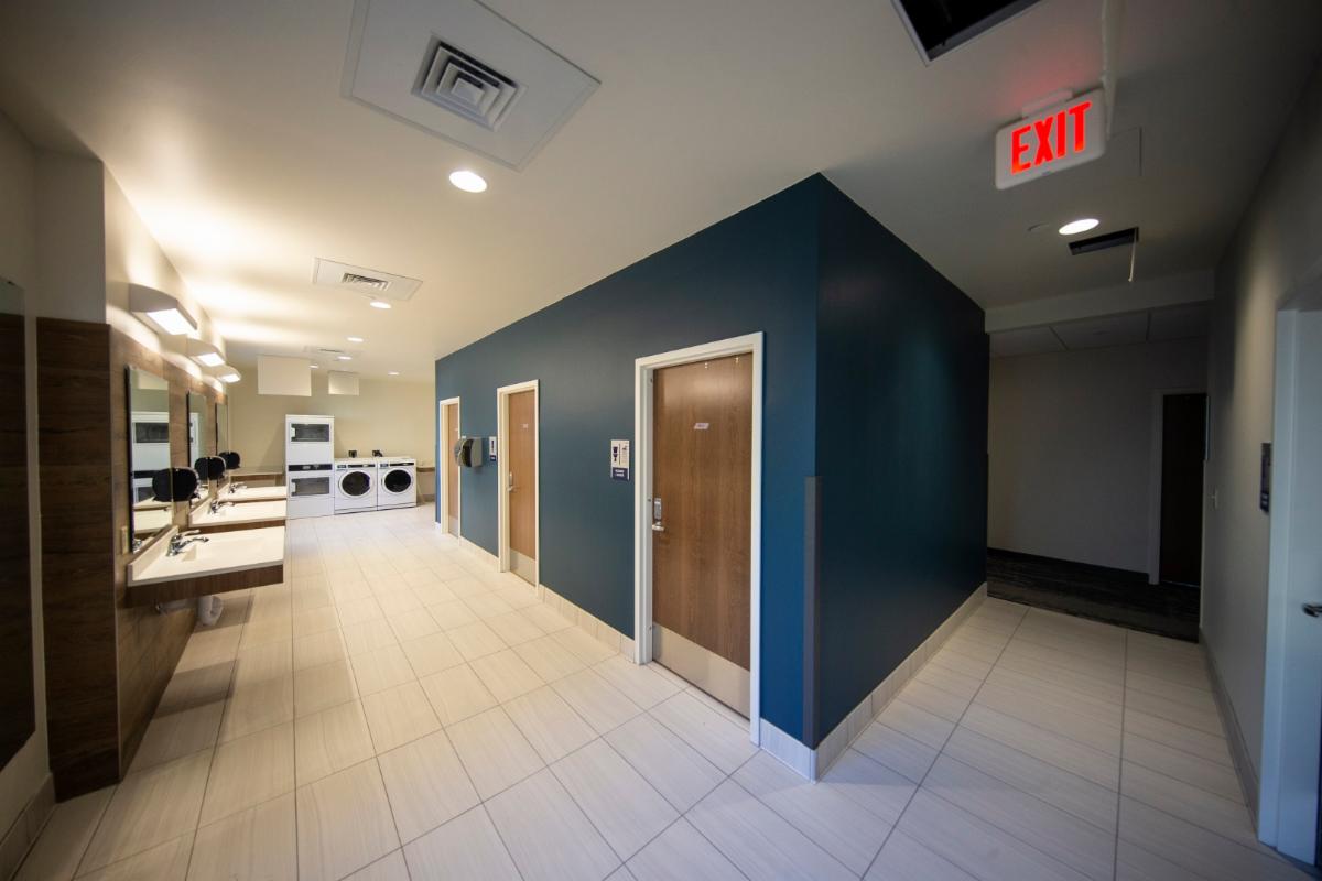 Photo of a bathroom in Gold Hall. Blue walls and white tile floors. Sinks and washers and dryers are visible. Wooden doors on the interior blue wall lead to individual toilet and shower rooms.