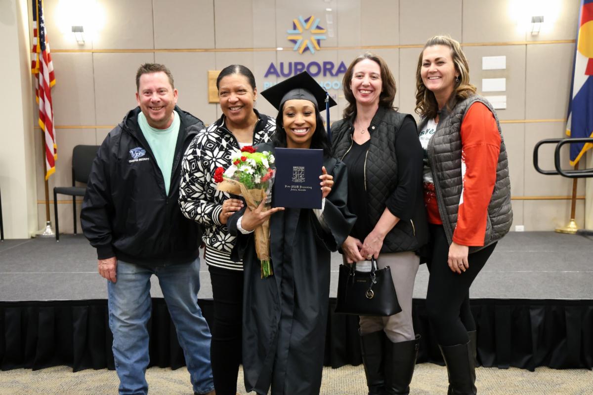 APS graduate holding her diploma and a bouquet of flowers with parents and staff members around her.