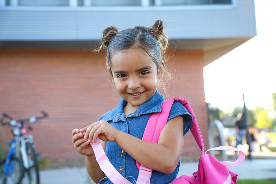 Smiling Elementary school student standing outside of school carrying a pink backpack.