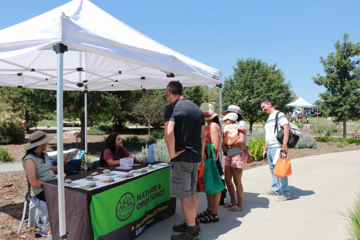 This image of people standing at a tent talking with people behind a table links to a webpage with more information about this event