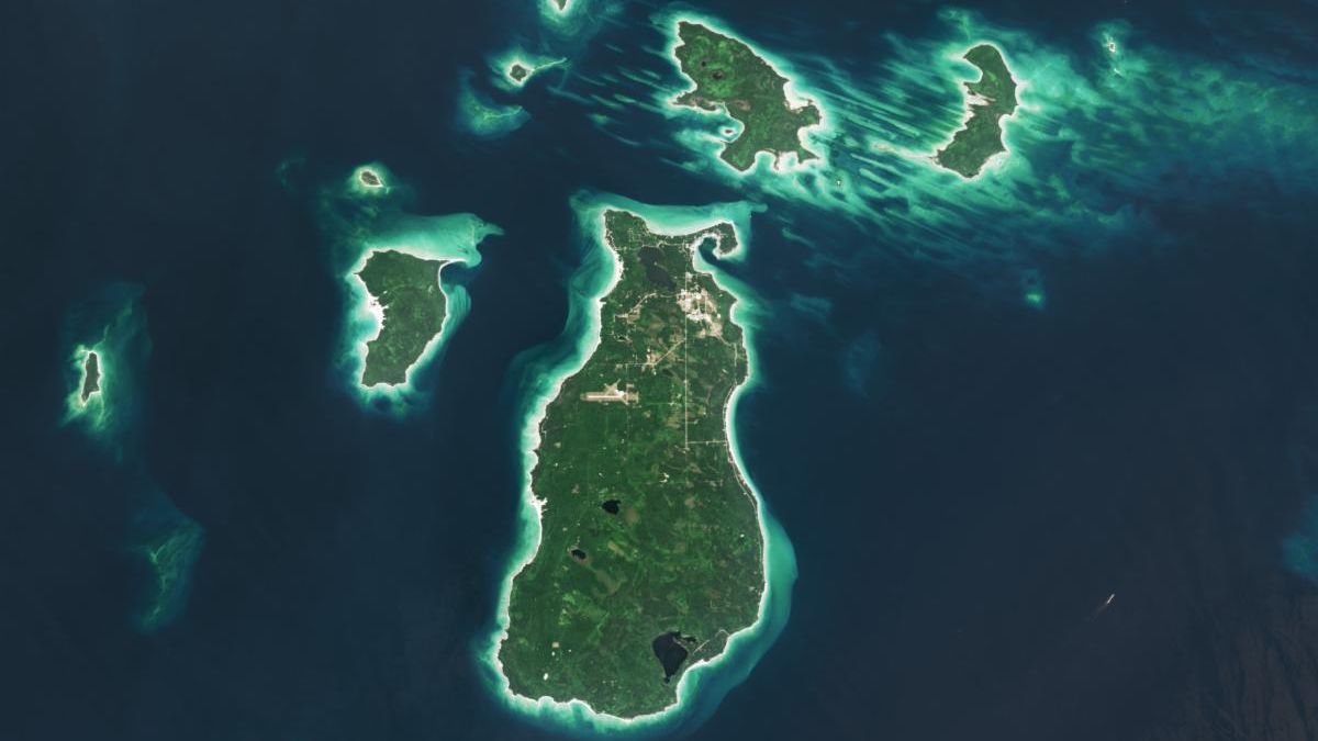 A satellite image shows several islands in Lake Michigan. The largest one, Beaver Island, is in the center. The islands are mostly green and vegetated, with bright sandy areas on their perimeters. Shallow waters near the land appear turquoise, and deeper waters are dark blue.