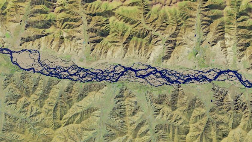 A false-color satellite image shows numerous intertwining channels of blue water spanning the middle of the scene from left to right. The river is bordered on the top and bottom by mountainous terrain, which appears tan and light green.