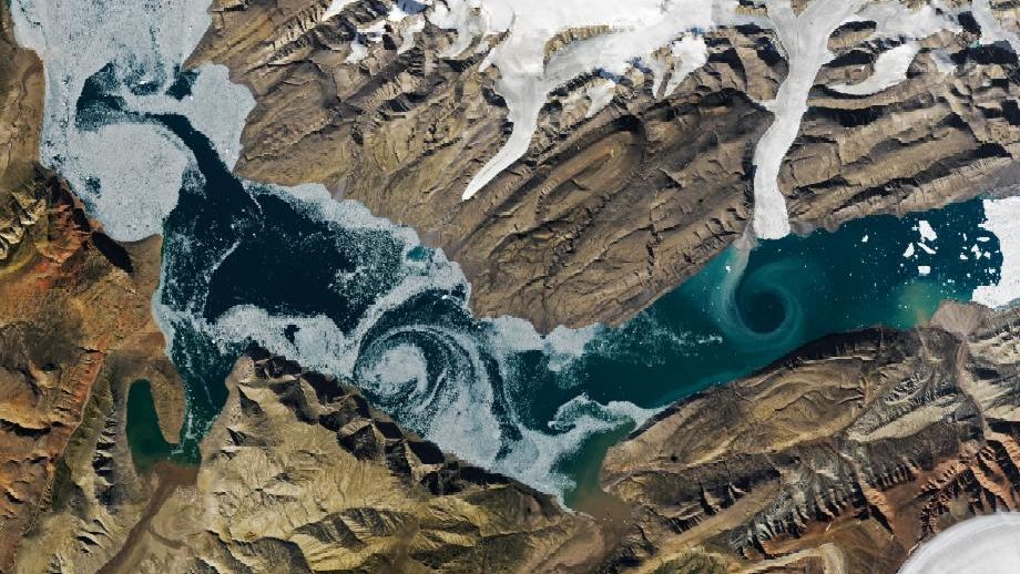 A V-shaped fjord cuts through barren brown land, with one patch of swirling water marked by white sea ice and another one colored turquoise by suspended sediment. Glacial ice flows into the fjord in several places.