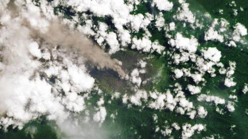 An ash-rich plume streams northwest from the volcano amidst scattered puffy, white clouds.