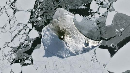A small ice- and snow-covered island with an active volcanic crater at its center is surrounded by drifting pieces of sea ice. A thermal signal and small plume appear in the crater, and ash darkens the snow on the volcano’s northern slopes.