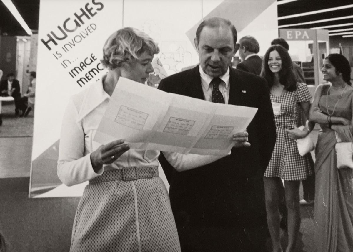 Virginia Norwood dressed in a white shirt and shirt shows a large piece of paper to a man dress in a dark suit.
