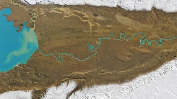 The outlet of bright blue Lago Argentino meanders across an otherwise dry, brown valley from left to right. The bottom third of the image is white, where snow covers higher elevations.