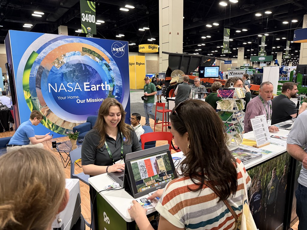 Allison Nussbaum stands behind a display table at a conference and is typing on a computer. On the monitor is the name "Monica" displayed in letters of Earth's surface. Attendees are gathered around the table. In the background is a large wall that reads "NASA Earth".