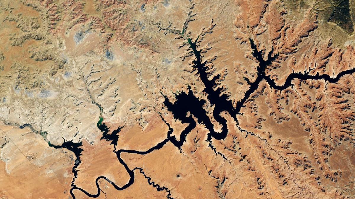 Water in Lake Powell is at record-low levels and looks like a wide river winding through a light orange desert landscape. The Glen Canyon Dam and the town of Page, Arizona, are visible in the lower left.
