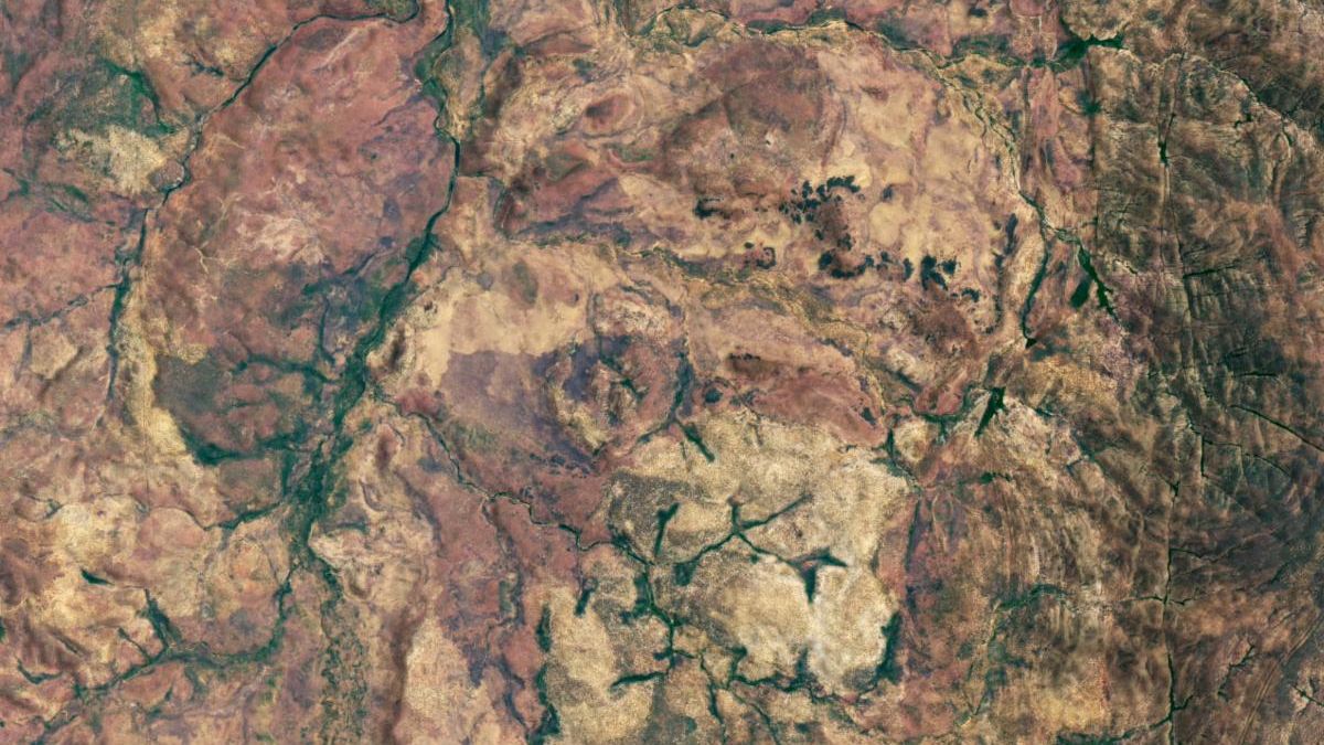 A satellite image shows the land surface of part of Australia’s Northern Territory. The image highlights the location of the Goyder impact structure, the eroded remnants of the central uplift portion of an impact crater. The central uplift remnants appear as small hills in the center of the image, with a light-colored ring of sandstone surrounding them. Most of the dry-looking surface is tan or red, but stream valleys are green.