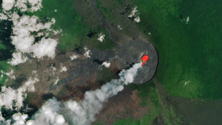 A satellite image shows the wide crater of Nyamulagira Volcano. Light brown to black lava flows radiate from the crater, and the rest of the volcano’s slopes are green with vegetation. An infrared signature shown in red indicates an area of heat within the crater, and a gray volcanic plume drifts from it to the southwest.
