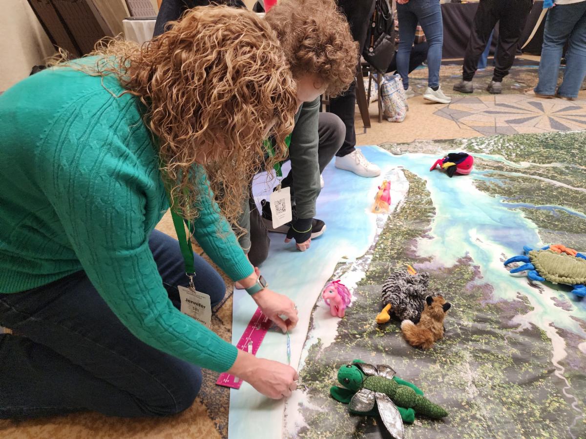 Two participants at the Maryland Association for Environmental and Outdoor Education (MAEOE) conference are gathered around the Chesapeake Bay mat (a large Landsat mosaic of the bay), using a paperclip chain and ruler to measure the length of Earth features such as Ocean City.