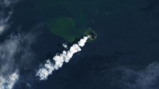 A natural-color Landsat image of the small volcanic island of Home Reef in January 2026 following an eruption. The volcanic plume appears as a long white trailing cloud and sediment in the water surrounding the island appears green.
