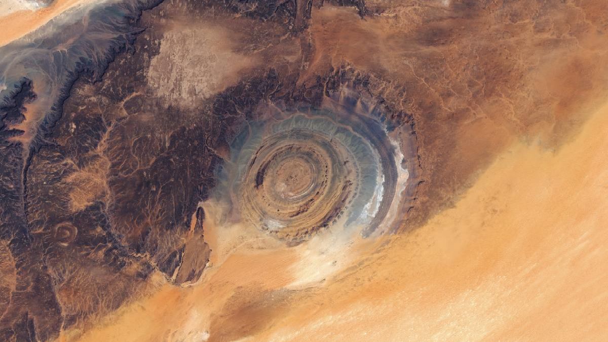 A large "bull's-eye" feature composed of concentric rock formations of various colors is imprinted on the edge of a dark plateau flanked by desert sands.