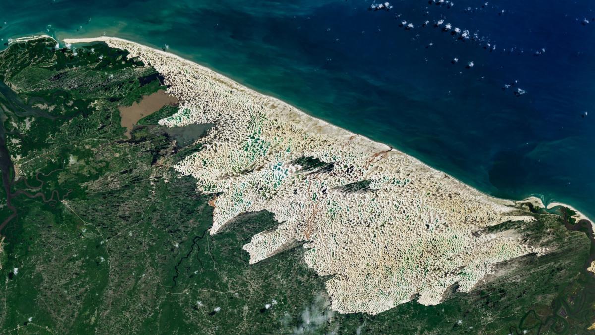 Bright white sand dunes stretch for dozens of kilometers between the dark blue Atlantic Ocean and green coastal vegetation. Blue and green freshwater lagoons fill low areas between the dunes, while dark-green vegetated patches lie within the dune field.