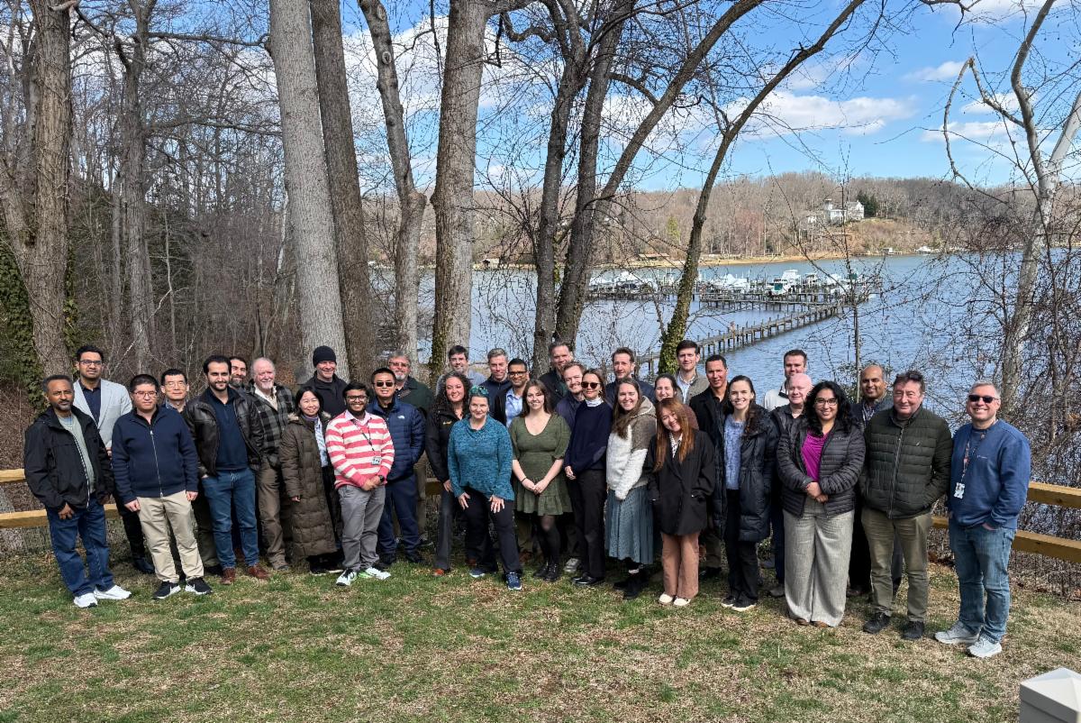 A group photo of the HLS science group at the face-to-face meeting. The group stands in front of a lake and several trees on a patch of green grass. 