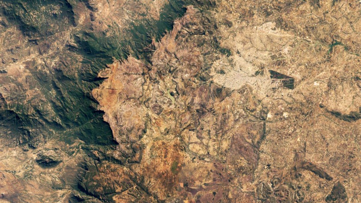 This satellite image centers on the serrated edge of the Huila plateau in Angola. The sheer cliff wall forms a rough C shape and is highlighted by dark shadows and a band of green vegetation. The lowlands to the left are tan-colored arid coastal plains, and the more-temperate plateau on the right side is greener.