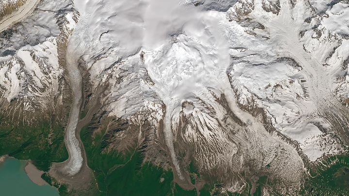 A detailed satellite image displays white, snowy mountains with Mount Spurr's volcanic summit in the center. The vent on Crater Peak is near the bottom of the image and has slightly less snow cover. The bottom-left is vegetated and green, with a blue-green lake's margin visible in the corner.