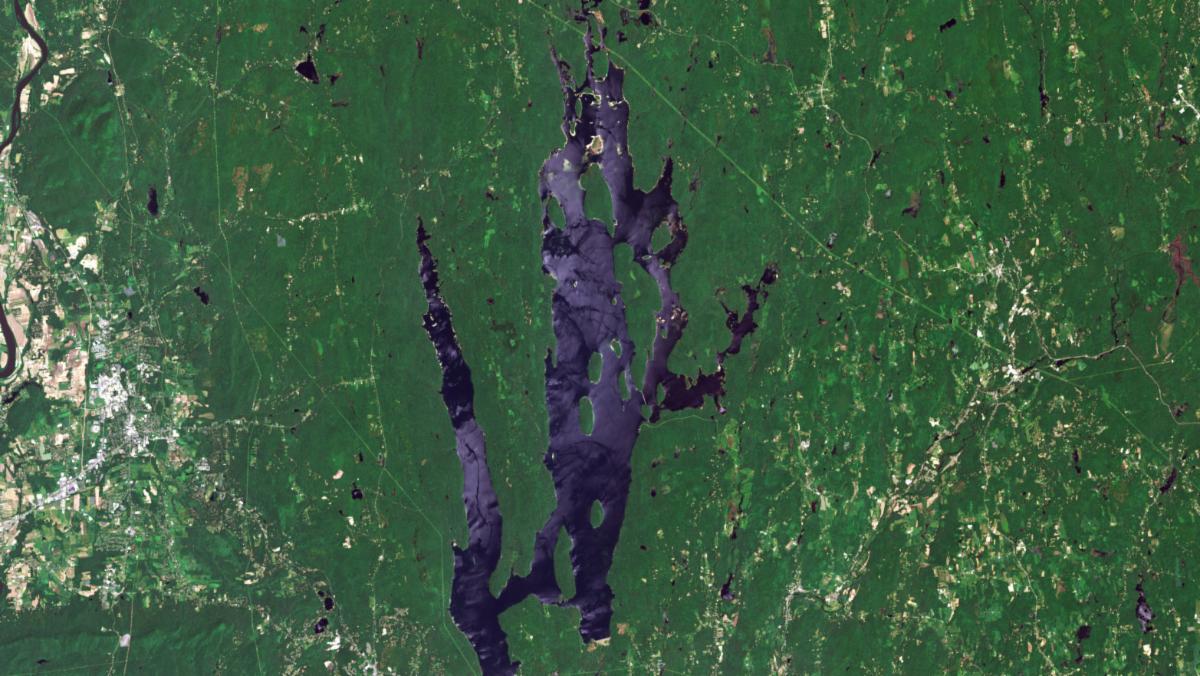 A satellite image shows the Quabbin Reservoir in central Massachusetts. The reservoir (blue) has an irregular shape that resembles a flooded river valley with multiple arms. The reservoir is surrounded by forest (green) and some towns and roads. A label for Dana Common shows the location of a town that was razed when the reservoir was created.
