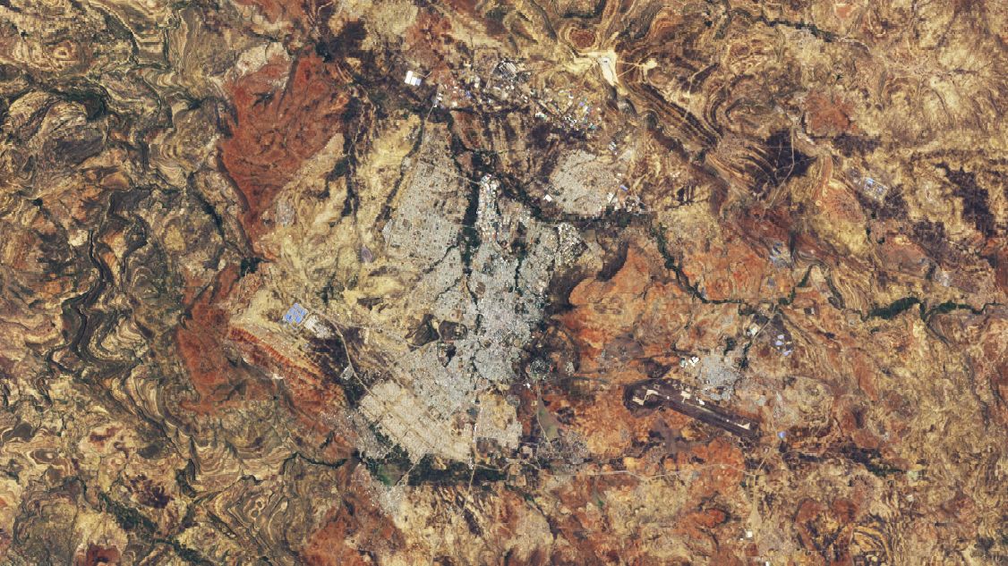 A Landsat image shows Mekele from above on May 31 2025. Now the city appears as a much larger gray urban area spreading across the center of the frame with labels marking planned development to the northwest unplanned development to the southwest an industrial park to the west and an airport with a visible runway to the southeast. The surrounding terrain shows rugged reddish-brown and tan hills with sparse vegetation.