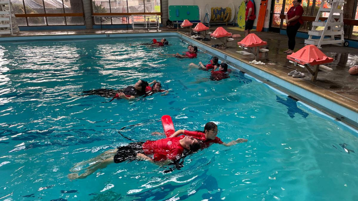 Participants wearing red flotation devices practice water safety skills in an indoor swimming pool while instructors observe from the pool deck. Colorful safety equipment is visible along the side of the pool.