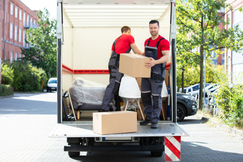 Two Young Men Unloading And Stacking The Brown Cardboard Boxes On Moving...