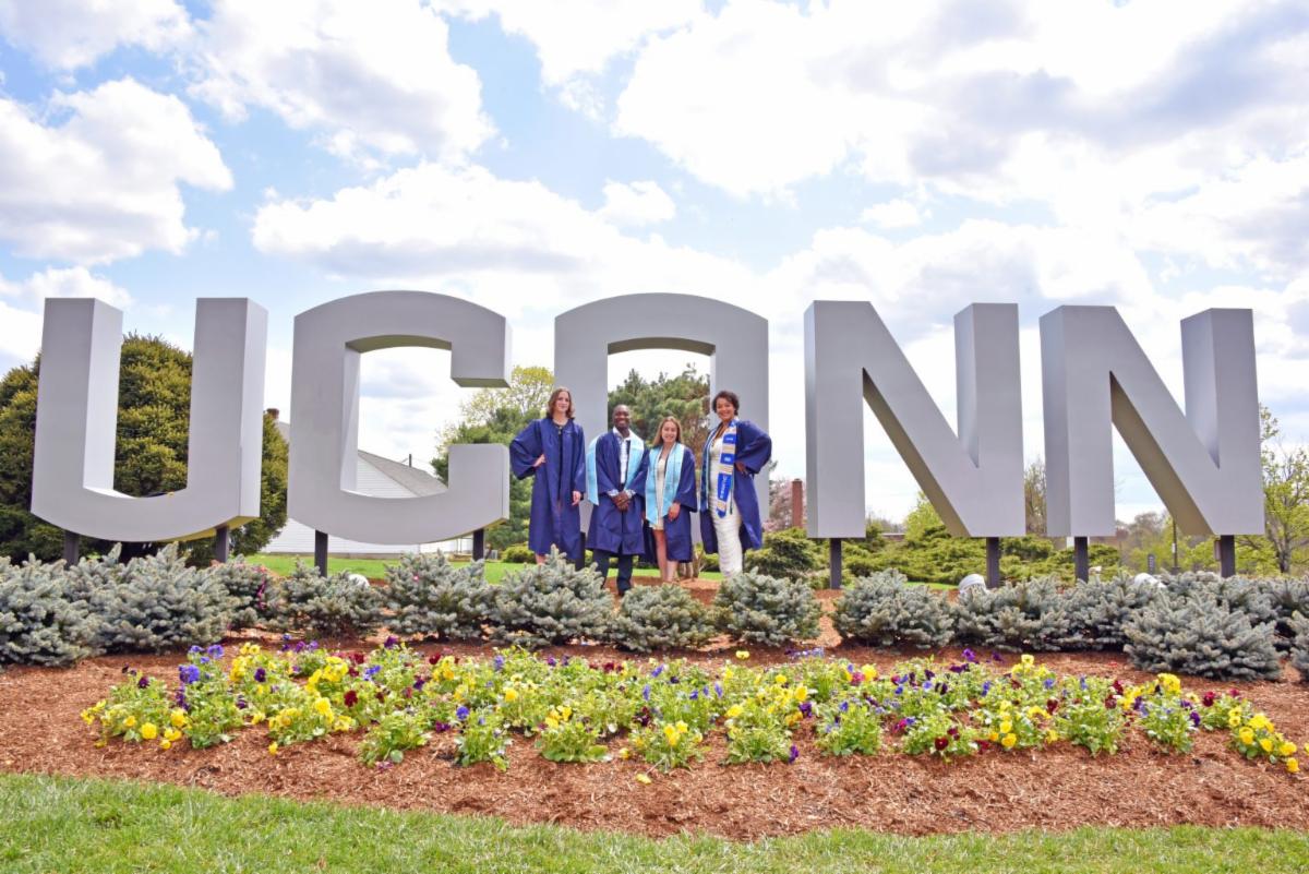 UConn grads posing in cap and gown in front of a UConn sign