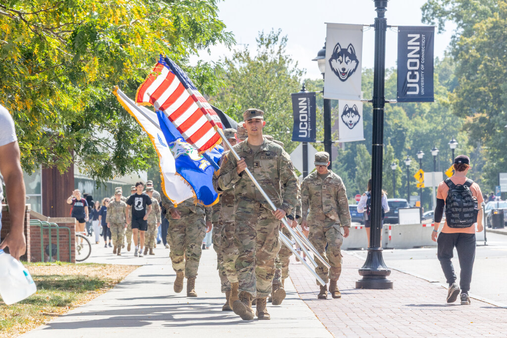 A group of US Army ROTC cadets in uniform march down a sidewalk on the University of Connecticut campus with one cadet carrying the American flag and another holding the Connecticut state flag 