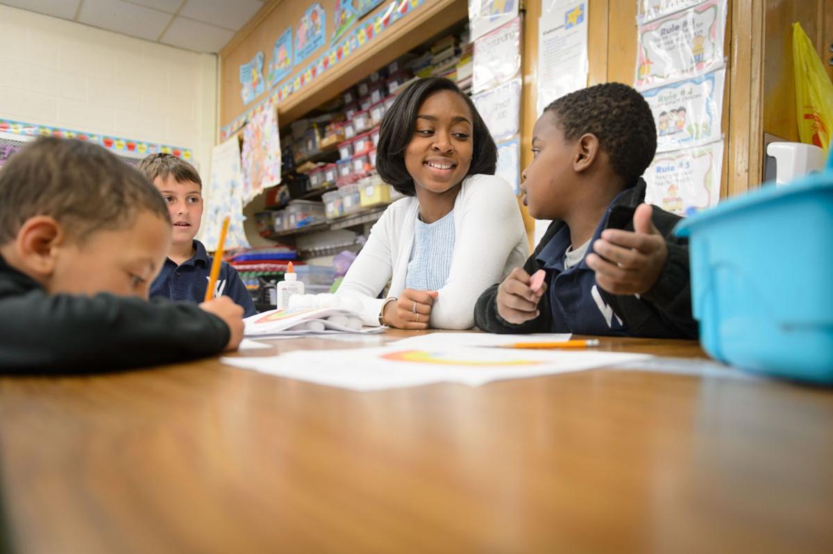 Picture of a teacher looking a her student.  Another student is writing, and another one looking at her.