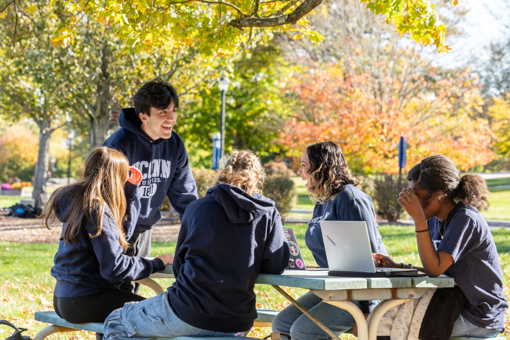 Group of students wearing UConn sweatshirts sitting on a bench. They are talking and have smiles on their faces. Fall foliage in the background.