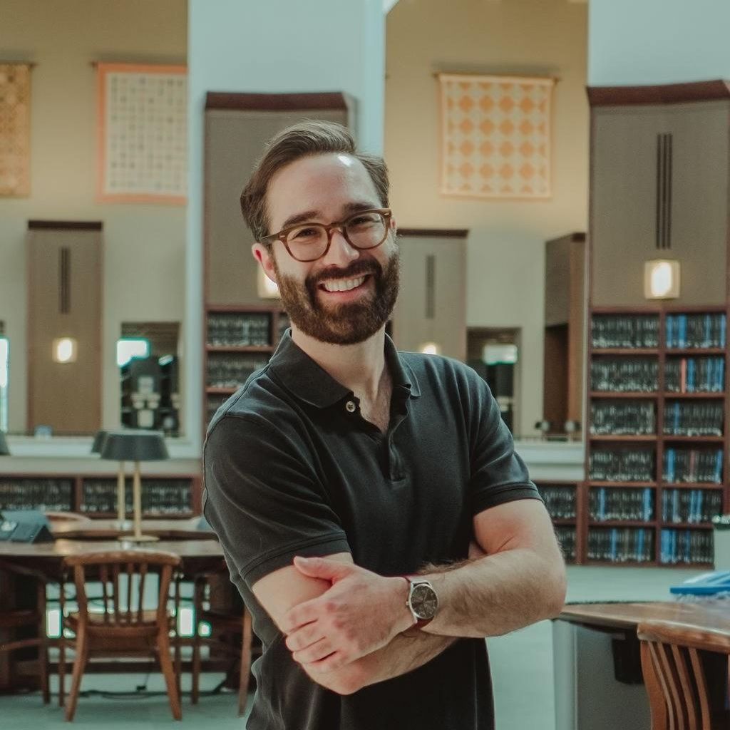Photo of Christopher Ciarcia with arms crossed at library with a shelf of books behind him. 