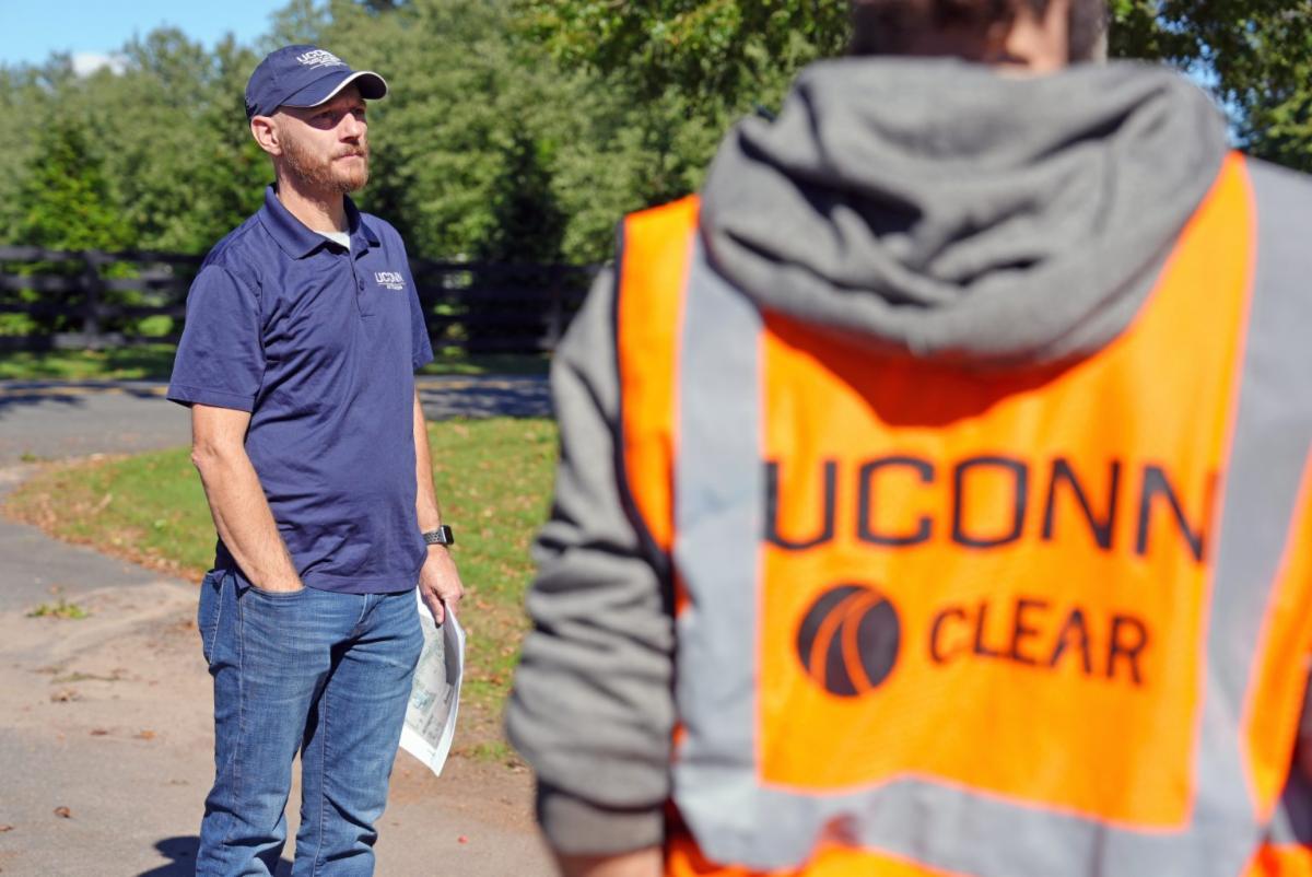 A gentleman standing with his hand on his pocket.  In front of him there is another gentleman wearing an orange UConn CLEAR sweatshirt.