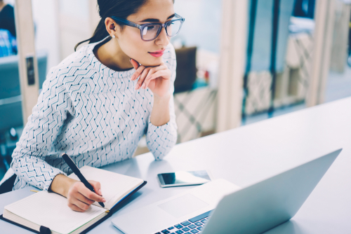 Pensive female administrative assistant checking report on laptop reading information and noting data_skilled woman watching online video online pondering on creating publication writing in notepad