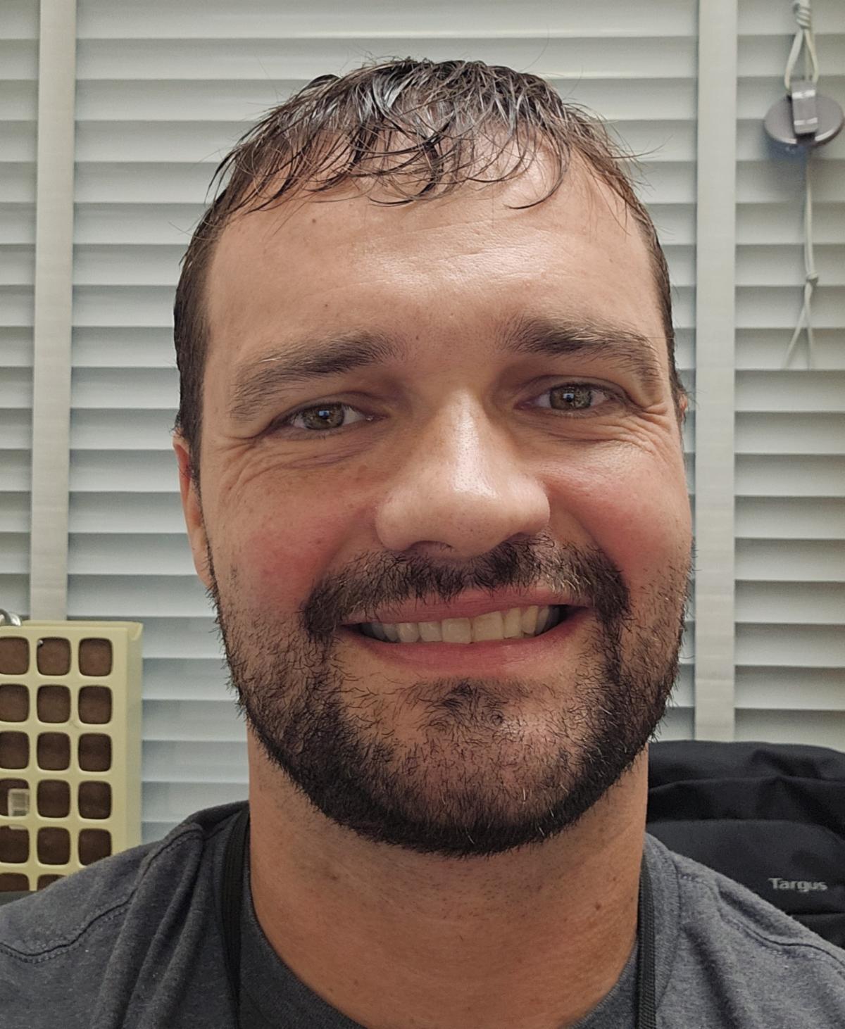 - Close up headshot of a slightly bearded white man smiling at the camera.